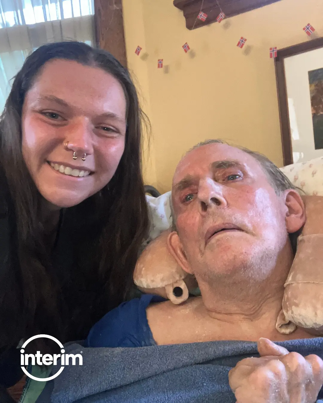 A hospice volunteer embraces an elderly person in a wheelchair beneath blooming trees, with the text “Become a Hospice Volunteer – Be a source of comfort in your own community.”
